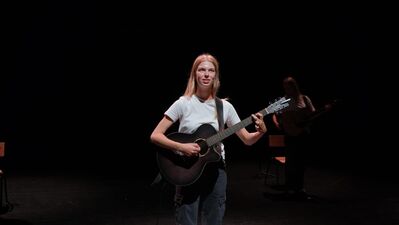Een jonge blonde vrouw staat in een wit t-shirt en met een gitaar op het podium.