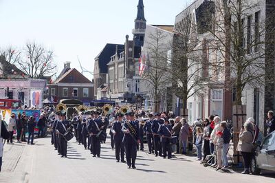Een fanfare loopt over een marktplein. Ze zijn omgeven door oude huizen en kerken.